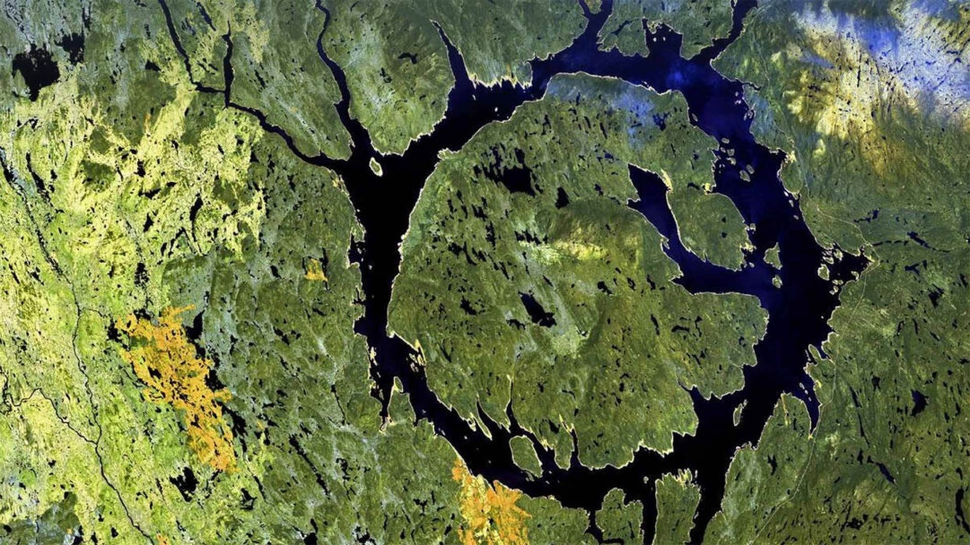 A breathtaking 4K wallpaper showcasing the vast, ring-shaped Manicouagan Crater in Quebec, Canada, as seen from an aerial Earth view. Its striking dark blue reservoir forms a dramatic circular pattern against the surrounding rich tapestry of green forests and scattered lighter terrain.