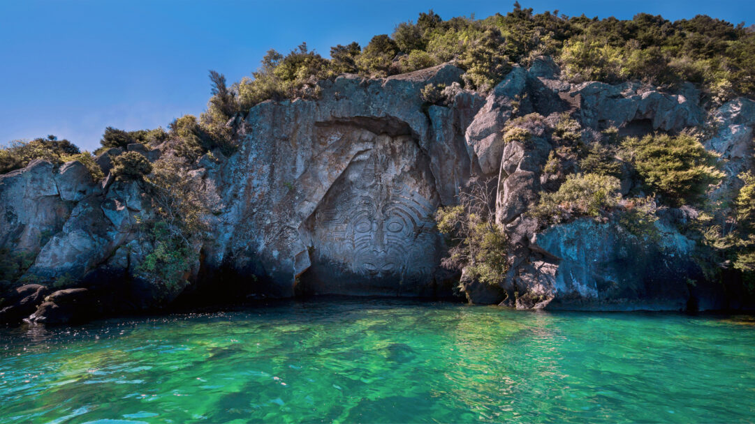 A captivating 4K wallpaper featuring the monumental Māori Rock Carving of Matahi Whakataka-brightwell, majestically situated on a rugged cliff face rising from the clear waters of Lake Taupo in New Zealand. Sunlight illuminates the carving's intricate details and the strikingly vibrant, emerald-green lake, creating a powerful sense of ancient artistry meeting serene natural beauty.