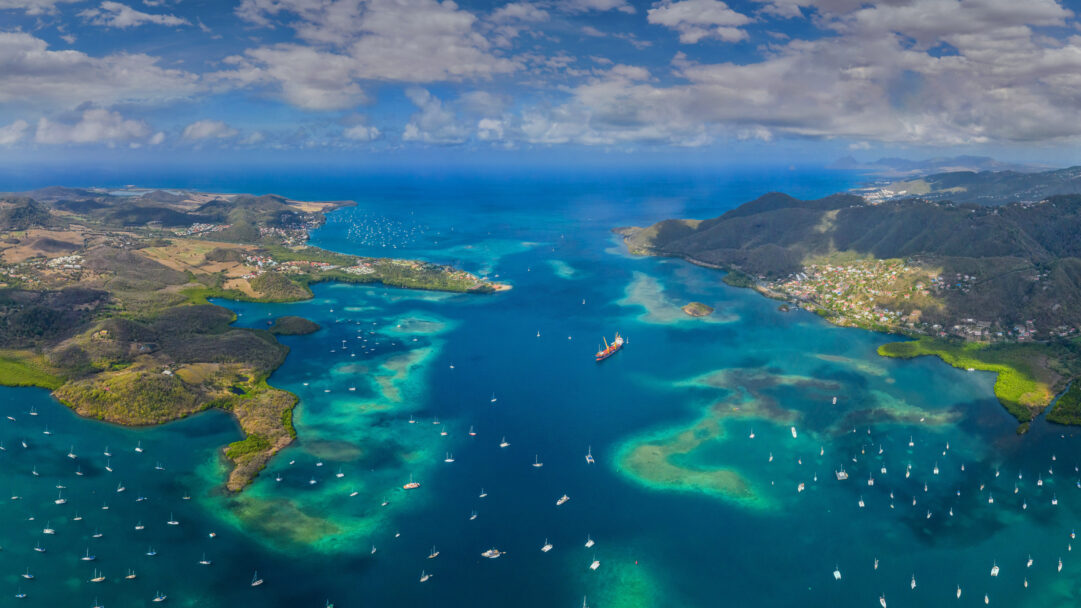 A breathtaking 4K wallpaper captures an aerial view of numerous boats dotting the crystal-clear turquoise waters of Martinique in the Lesser Antilles, Caribbean Sea. Sunlight beautifully illuminates the shallow coral reefs and deep blue channels, creating a mosaic of vivid colors against the lush green island and distant cloudy sky.