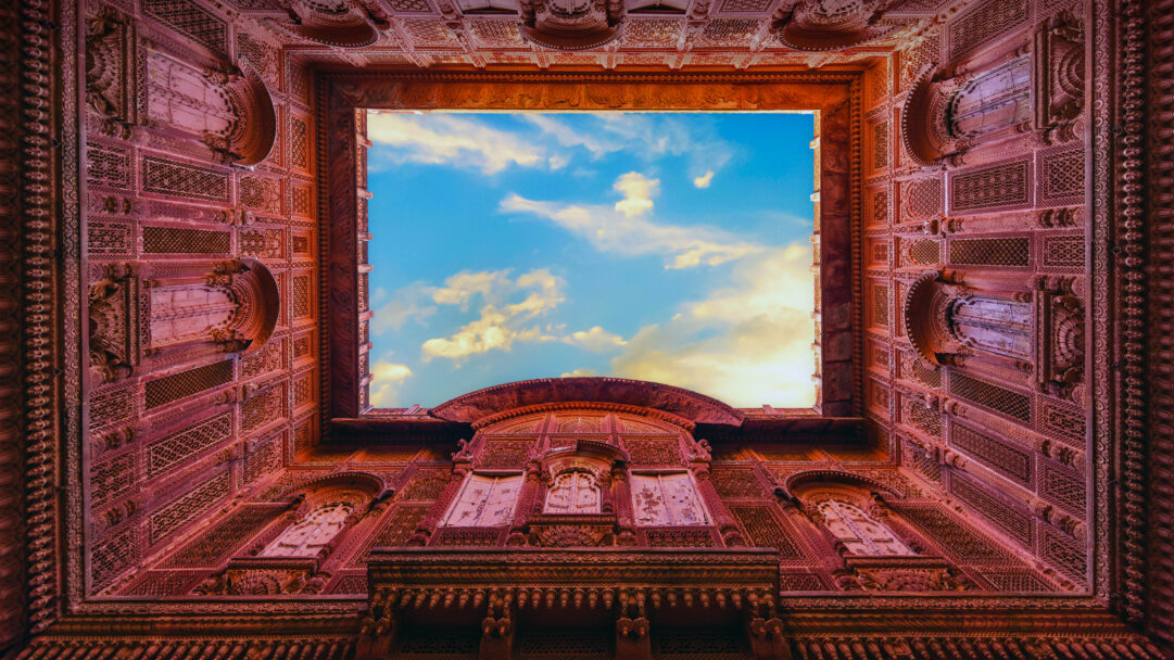 A magnificent 4K wallpaper showcases the intricate architecture of Mehrangarh Fort in Jodhpur, India, viewed from below as it frames a clear sky. The incredibly detailed red sandstone carvings line the square opening, with the serene blue sky and soft white clouds creating a striking contrast and a sense of timeless grandeur.