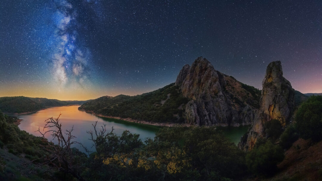A breathtaking 4K wallpaper captures the stunning Milky Way arching across the night sky above the winding Tagus River in Monfragüe National Park, Spain. The celestial river of stars illuminates the vast dark landscape, with soft orange and blue hues reflecting on the calm river surface, evoking a profound sense of natural wonder.