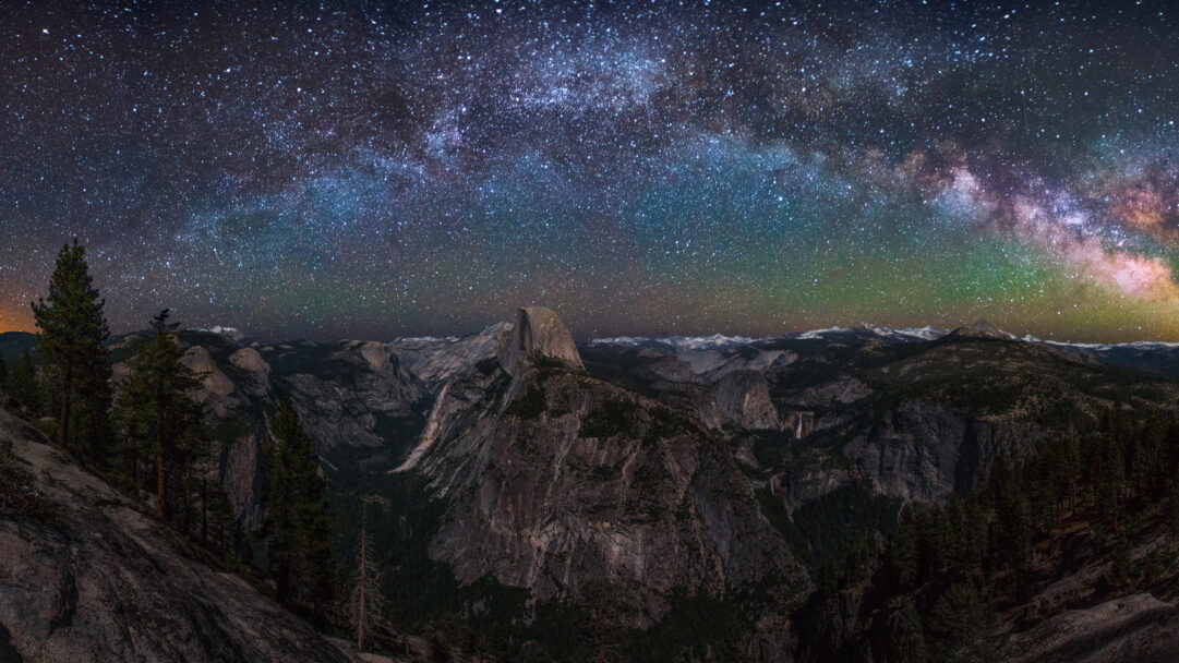 An awe-inspiring 4K wallpaper captures the Milky Way rising majestically above Half Dome in Yosemite National Park. The vast, star-filled expanse of the galaxy glows with vibrant blues, purples, and greens, creating a breathtaking celestial canopy over the dramatically lit granite cliffs and dark forest below.