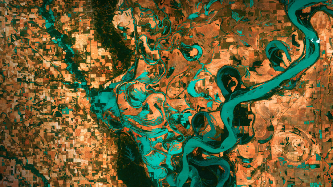 A breathtaking 4K wallpaper of an aerial view reveals the Mississippi River winding along the Arkansas-Mississippi border, flanked by vast agricultural fields and segments of dark forest. The vibrant teal of the braided river and its numerous oxbow lakes dramatically contrasts with the grid-like patterns of the rust-orange cultivated land, creating an abstract and dynamic landscape.