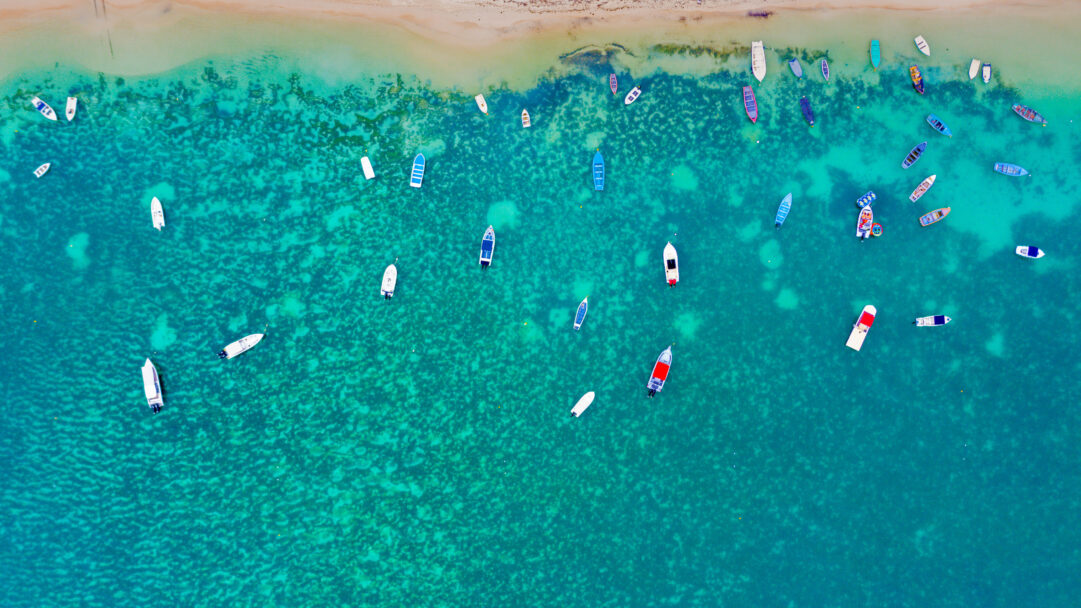A breathtaking 4K wallpaper showcasing an aerial view of Mont Choisy Beach in Mauritius, dotted with numerous colorful boats. The crystal-clear turquoise water reveals the vibrant coral patterns beneath, creating a captivating contrast with the varied shapes and hues of the anchored vessels.