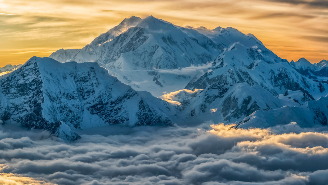 A breathtaking 4K wallpaper showcasing Mount Logan and other majestic snow peaks in Kluane National Park, Yukon, at sunset. The golden hues of the sunset illuminate dramatic clouds that swirl around and below the towering, snow-capped mountains, creating a serene and powerful mood.