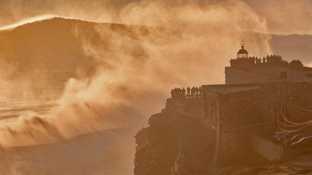 An awe-inspiring 4K wallpaper showcases the Nazaré Lighthouse and Forte de São Miguel Arcanjo in Portugal, perched on cliffs overlooking immense, breaking waves. The colossal waves erupt into a golden cloud of sea spray, backlit by a warm, ethereal light, creating a powerful natural spectacle that captivates onlookers.