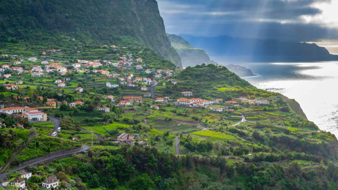 A majestic 4K wallpaper showcases a vibrant village built into the dramatically steep, terraced green hillsides of Madeira, Portugal’s North Coast. Sunlight dramatically breaks through heavy clouds to illuminate a shimmering path on the dark ocean waters, contrasting beautifully with the rich, deep green of the surrounding landscape.