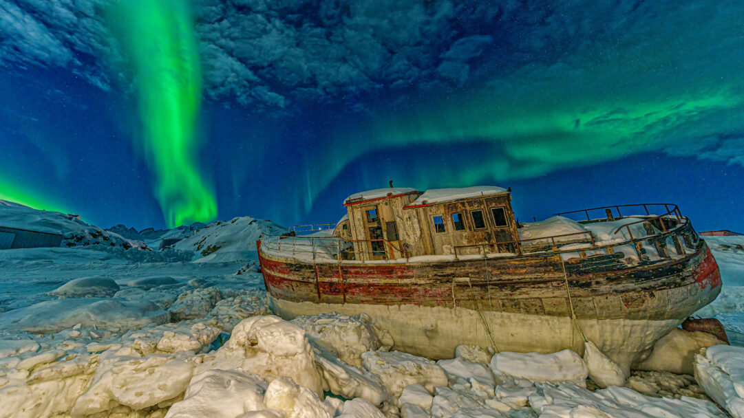 A magical 4K wallpaper of the Northern Lights dancing over a stranded, weathered boat in the icy grip of a Tasiilaq, Greenland winter. Vibrant green aurora streaks illuminate the star-dusted night sky, casting an ethereal glow over the snow-covered wreckage and frozen landscape, evoking a sense of majestic solitude.