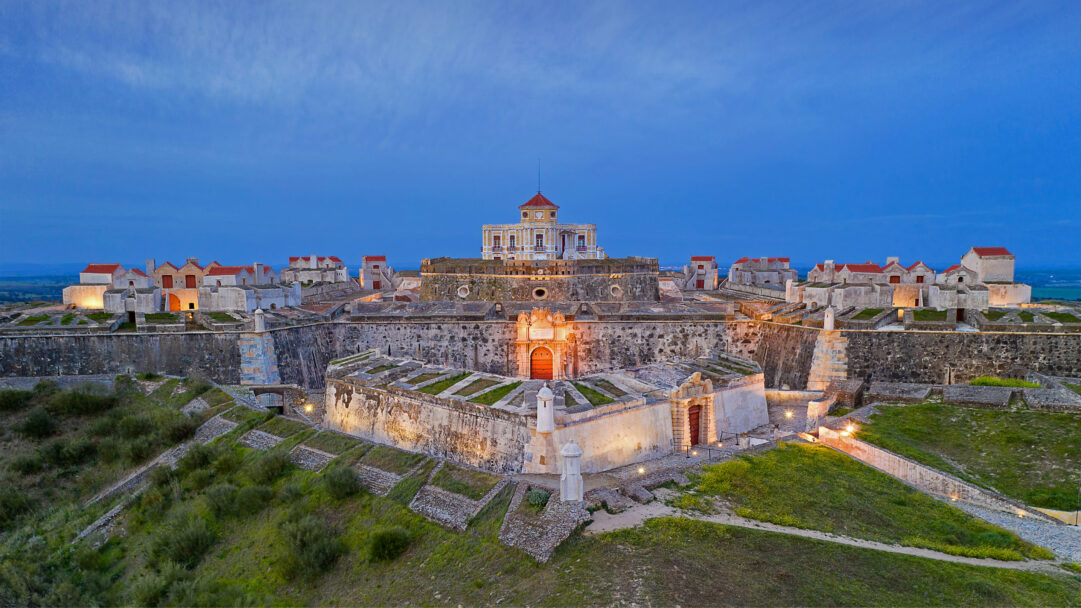 A captivating 4K wallpaper showcases the grand Nossa Senhora da Graça Fort in Elvas, Portugal, a sprawling military fortification atop a hill under a twilight sky. Warm interior lights and strategic exterior illuminations cast a golden glow on the fort's ancient stone walls, contrasting dramatically with the deep, inky blue of the dusk sky above.