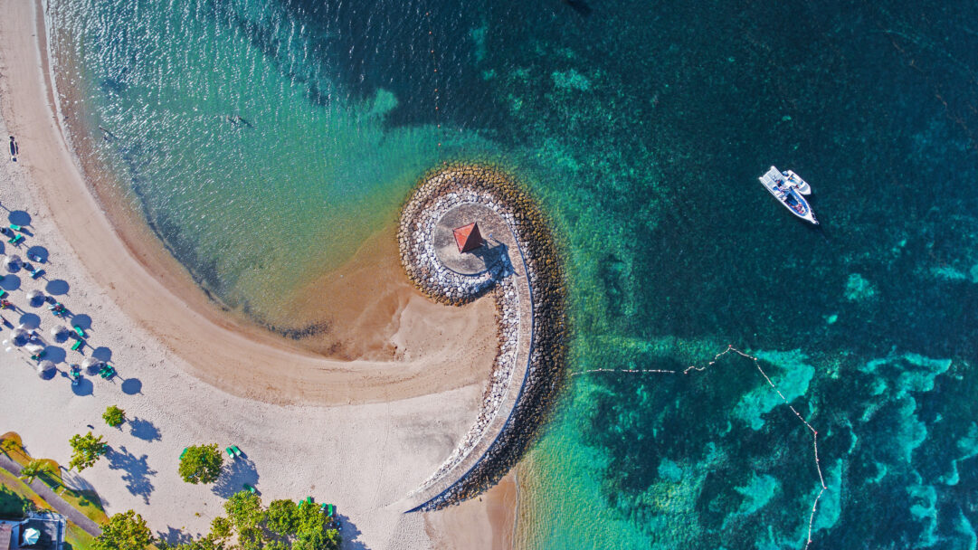 A serene 4K wallpaper depicting an aerial view of the Nusa Dua Coast in Bali, Indonesia, showcasing a pristine sandy breakwater beach and its distinctive curved stone barrier. The ocean displays a stunning gradient of shimmering turquoise and deep blue, revealing clear underwater details where a white boat peacefully rests.