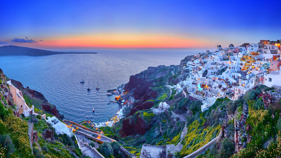 An enchanting 4K wallpaper capturing the panoramic Oia Village in Santorini, Greece, nestled on a cliff overlooking the calm Aegean Sea. The sunset paints the horizon in hues of orange and pink, beautifully complementing the deep blue sky and the soft glow of the village lights across the caldera.