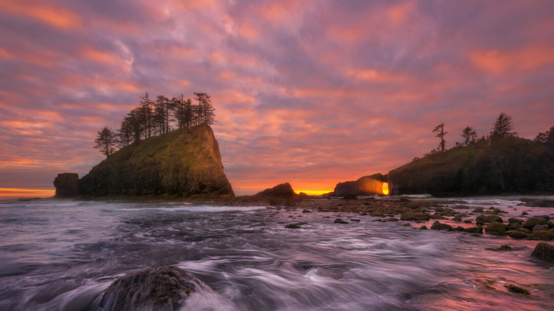 A captivating 4K wallpaper captures the Olympic Coast National Marine Sanctuary at sunset, featuring prominent sea stacks and a striking natural rock arch over the ocean. The setting sun dramatically illuminates the arch from within with a fiery glow, while vibrant orange, pink, and purple clouds reflect in the smooth, motion-blurred ocean waters below.