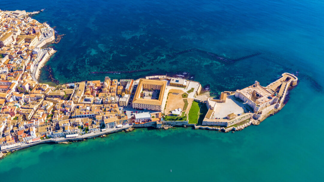 A breathtaking 4K wallpaper showcasing an aerial cityscape of Ortygia Island in Syracuse, Sicily, Italy, densely packed with historical buildings on a sun-drenched peninsula. The brilliant azure and emerald waters surrounding the island beautifully highlight its ancient fortifications and the warm hues of its terracotta rooftops, evoking a tranquil Mediterranean charm.
