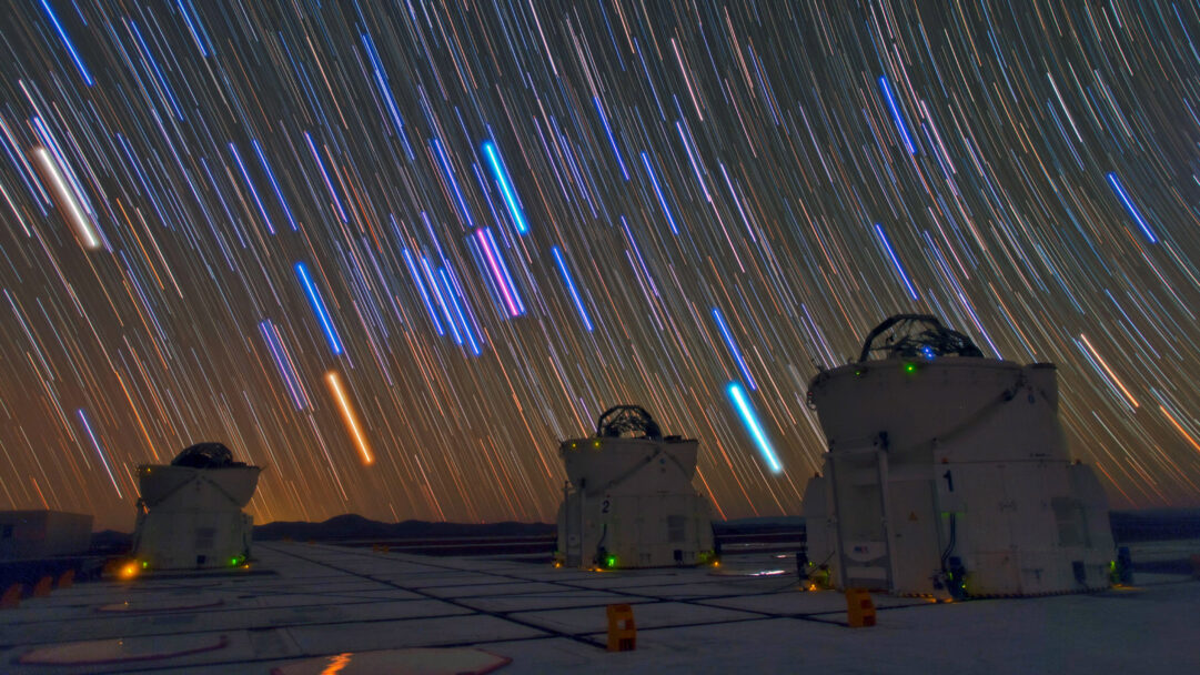 A celestial 4K wallpaper of the Paranal Observatory's telescopes standing resolute against a breathtaking night sky. The dark desert landscape is dramatically illuminated by countless vibrant star trails, streaking across the heavens in brilliant arcs of blue, white, and orange, conveying the Earth's majestic rotation.