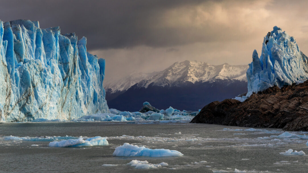 An awe-inspiring 4K wallpaper of the monumental Perito Moreno Glacier in Patagonia National Park, its massive blue ice wall towering over water littered with icebergs and distant snow-capped mountains. The glacier's vibrant, intricate blue ice is dramatically illuminated against a backdrop of dark, snow-capped peaks and a moody sky, emphasizing the raw, majestic power of this Patagonian landscape.