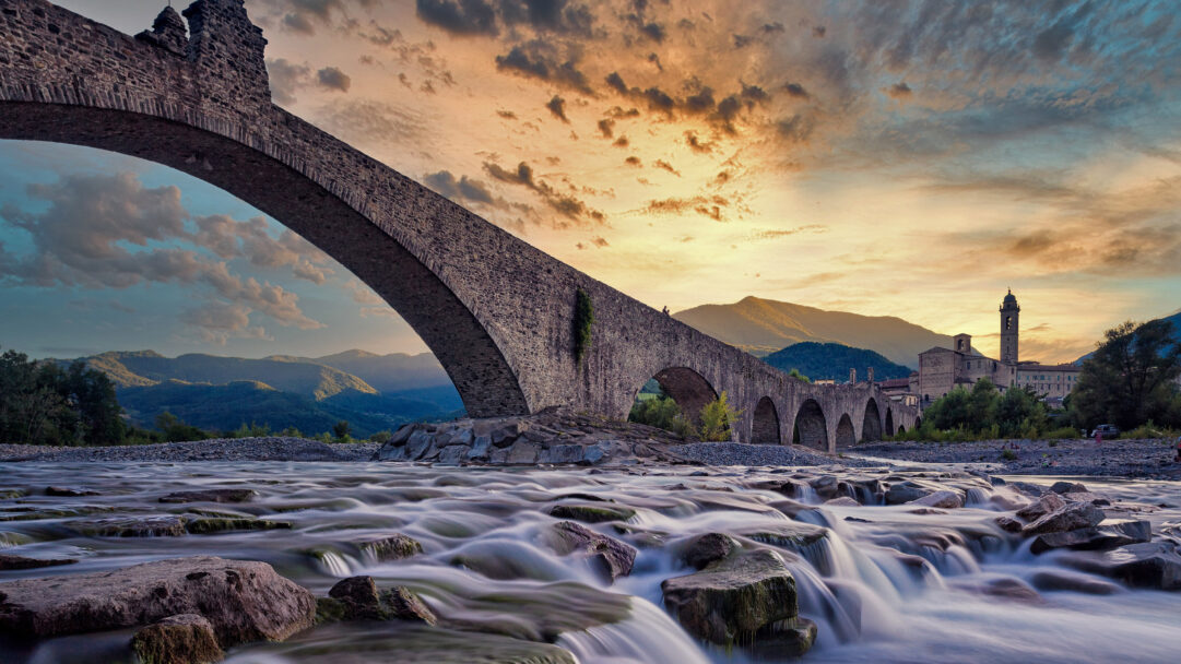 A magnificent 4K wallpaper showcasing the ancient Ponte Vecchio Bridge spanning a wide river in Bobbio, Italy, with distant mountains and a town nestled under a sunset sky. The long-exposure river water cascades over rocks, beautifully reflecting the orange and purple twilight, evoking a serene and timeless landscape.