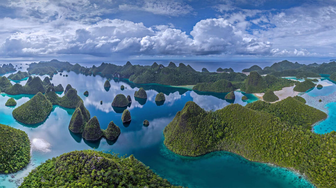 A breathtaking 4K wallpaper showcases an aerial view of the Raja Ampat Archipelago in Indonesia, where countless verdant, conical islands dot tranquil turquoise and deep blue waters. The calm sea magnificently reflects the dramatic white clouds above and the lush green of the islands, creating a stunning interplay of color and light that evokes a sense of pristine natural wonder.