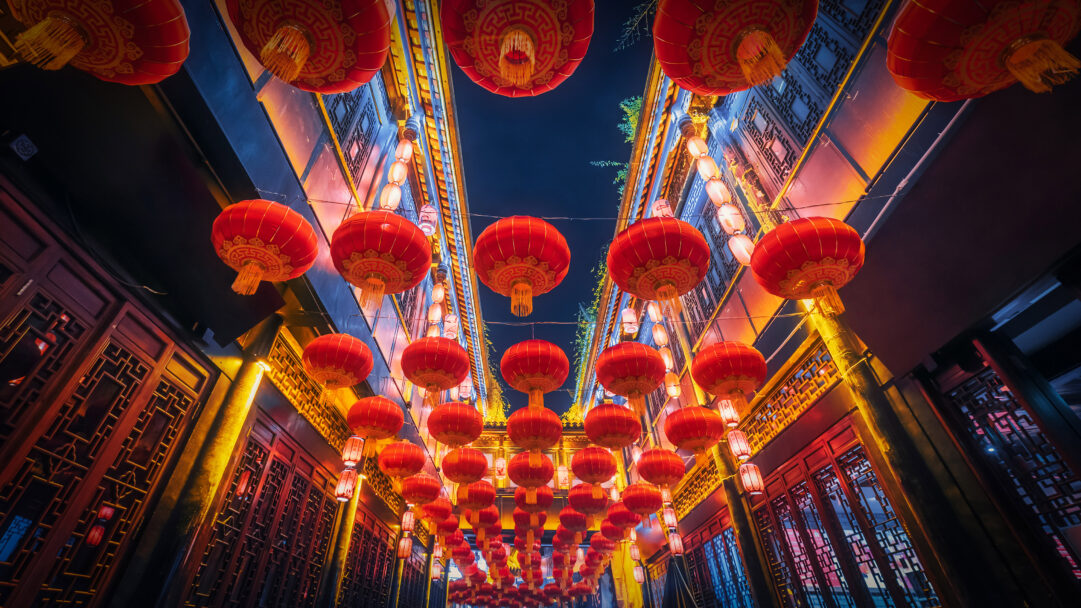 A mesmerizing 4K wallpaper showcases a vibrant night scene on Jinli Street in Chengdu, China, filled with countless glowing red lanterns. These ornate lanterns hang overhead, casting a warm, festive light that dramatically contrasts with the deep blue night sky and illuminates the traditional dark wood architecture of the street.