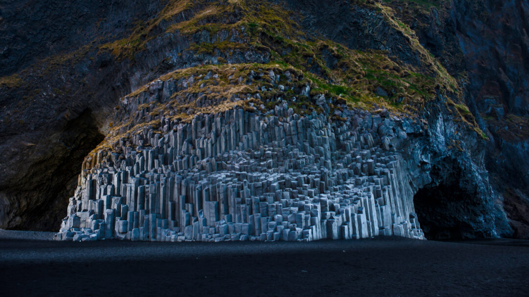 A captivating 4K wallpaper presents the iconic basalt columns rising from Reynisfjara Black Sand Beach in Iceland, nestled against a towering cliff face. The dramatic hexagonal pillars, glowing faintly with a cool grey light, contrast sharply with the deep, dark volcanic sand and the textured, moss-draped cliff above, creating a powerful and ancient geological tableau.