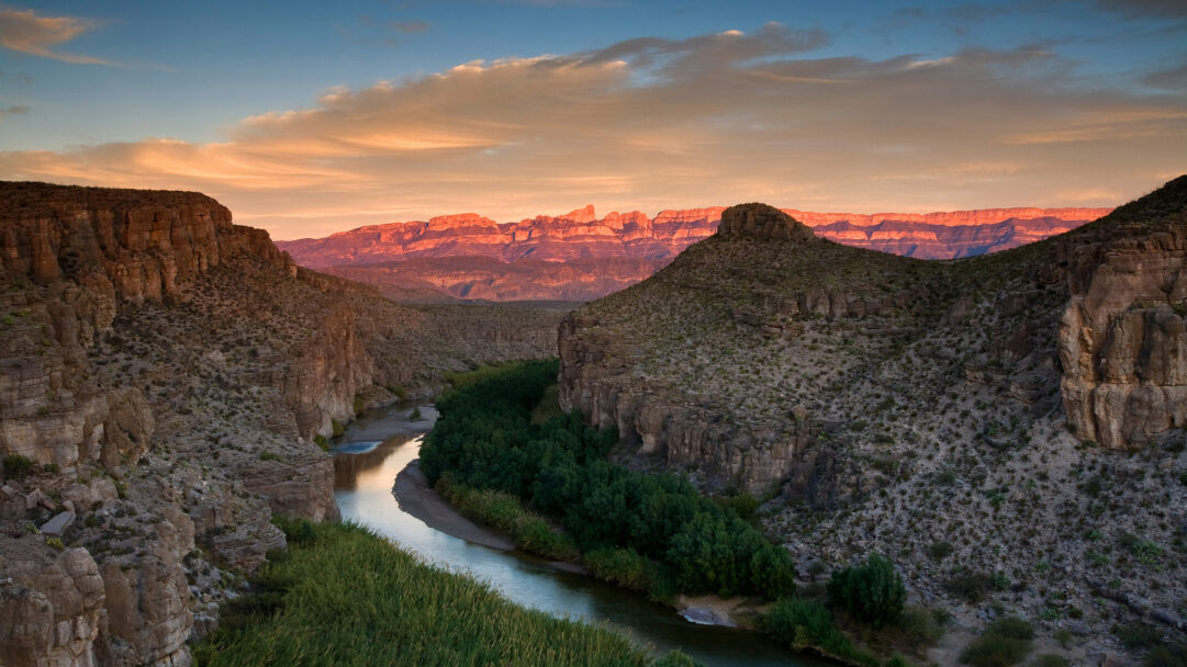 A majestic 4K wallpaper capturing a sweeping sunset view of the Rio Grande winding through a deep canyon in Big Bend National Park, Texas. Golden orange clouds streak across the sky, illuminating the rugged mountain range with warm, fiery hues that reflect softly on the calm river.