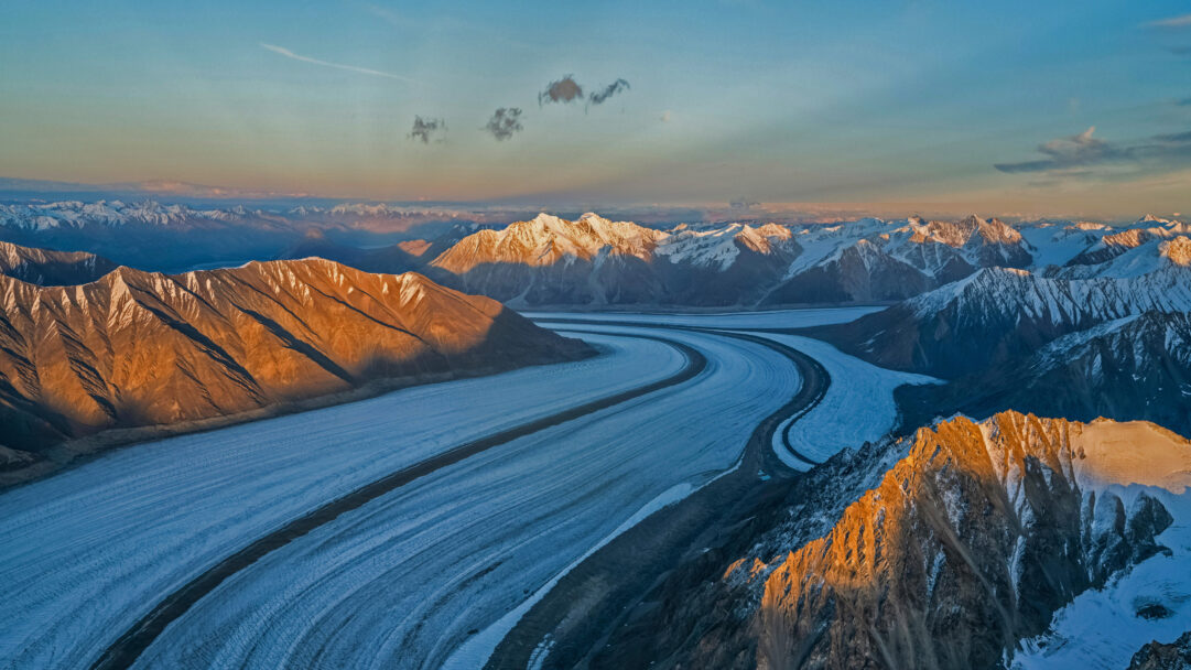 A majestic 4K wallpaper presents the expansive Kaskawulsh Glacier winding through the rugged peaks of the Saint Elias Mountains within Kluane National Park. Golden light illuminates the sharp, snow-dusted ridges of the surrounding mountains, dramatically contrasting with the deep blue ice of the glacier below, evoking a sense of ancient, untouched grandeur.