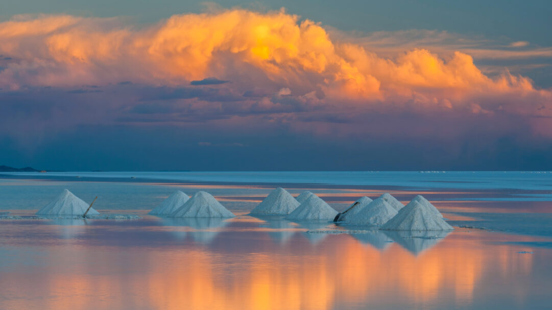 A breathtaking 4K wallpaper of Salar de Uyuni in Bolivia, featuring numerous glistening white salt cones resting on the shallow, reflective salt flat. Warm orange and golden clouds of the sunset intensely reflect across the water-covered salt flats, mirroring the vibrant sky and creating a stunning interplay of color and light.
