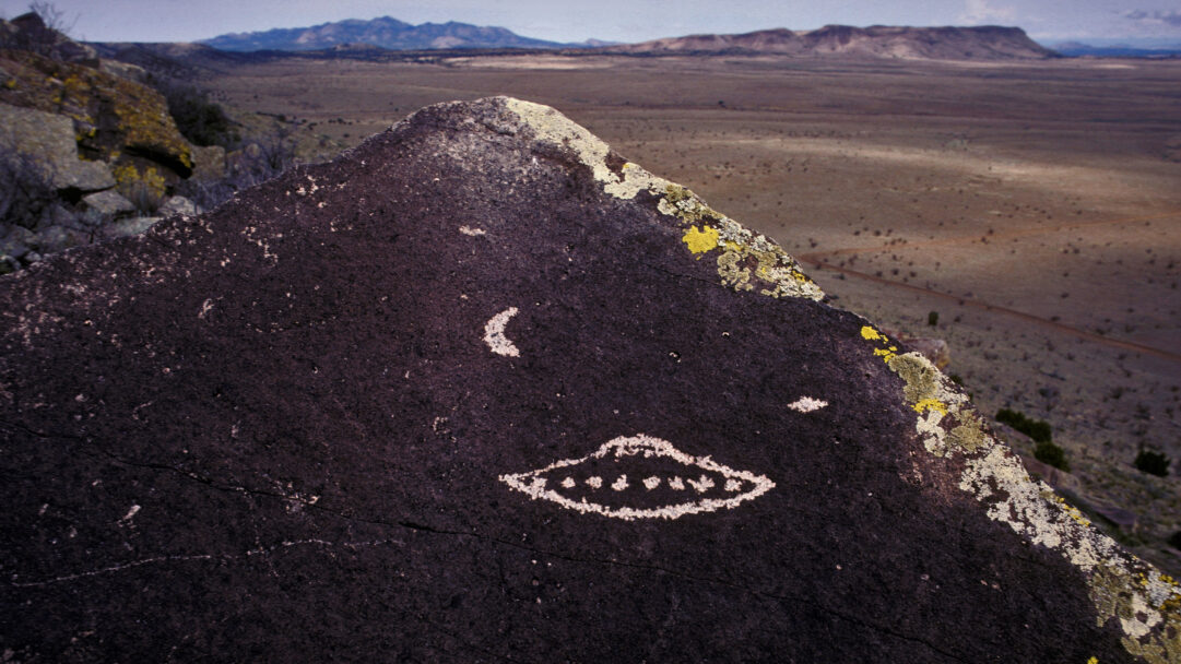 A mysterious 4K wallpaper features ancient Santa Fe, New Mexico rock art: a UFO petroglyph etched onto a dark, lichen-covered boulder overlooking a vast desert. The stark white of the stylized UFO, flanked by crescent shapes, creates an otherworldly focal point against the deep rock, setting a mood of ancient enigma across the expansive, muted landscape.