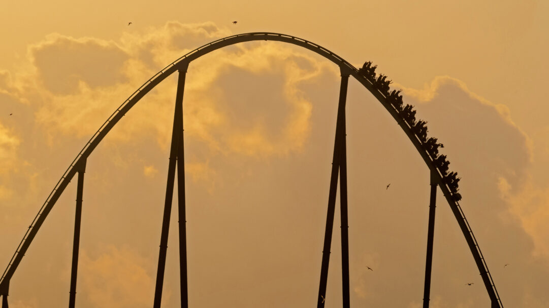 A breathtaking 4K wallpaper captures the majestic silhouette of the Shambhala Roller Coaster at PortAventura Park, ascending its highest peak during sunset. The track's dark outline and the riders' silhouetted, cheering forms create a powerful sense of height and exhilaration against the sky's dramatic golden-orange glow.