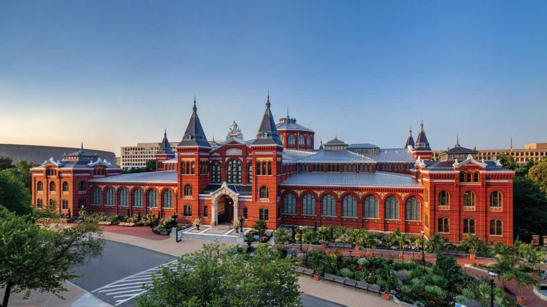 An iconic 4K wallpaper of the Smithsonian Arts and Industries Building, a historic landmark in Washington DC, presented majestically from an elevated view within its verdant urban setting. Its striking red brick facade and pale gray metal roofs are bathed in the golden hour's gentle light under a vast, cloudless blue sky, emphasizing its ornate Victorian details and serene grandeur.