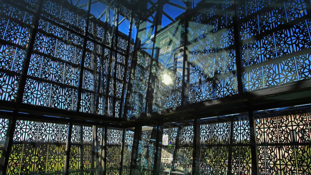 A captivating 4K wallpaper showcasing the distinctive facade pattern of the Smithsonian National Museum of African American History, seen against a bright blue sky and glimpses of green foliage. The dark, intricate geometric lattice is dramatically illuminated by a bright sunburst, creating dynamic patterns of light and shadow across the reflective surfaces and deep blue sky.