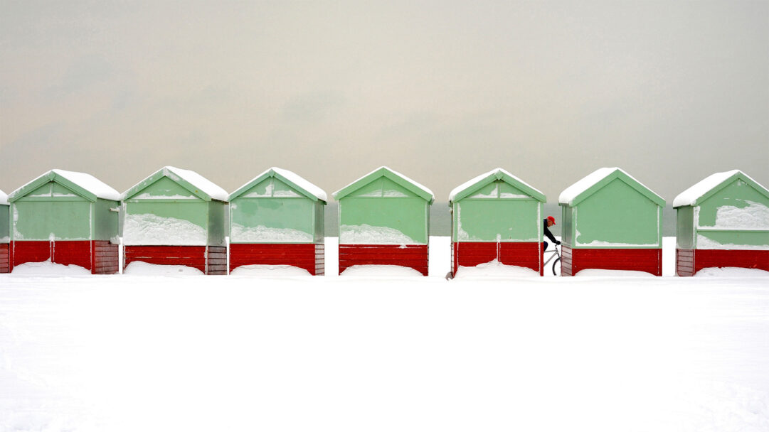 A minimalist 4K wallpaper presents a striking line of snow-covered beach huts on a winter's day in Brighton, England. The vibrant red and pale green huts stand out against the pristine white snow and vast muted sky, creating a serene, almost painterly, landscape as a person cycles by.