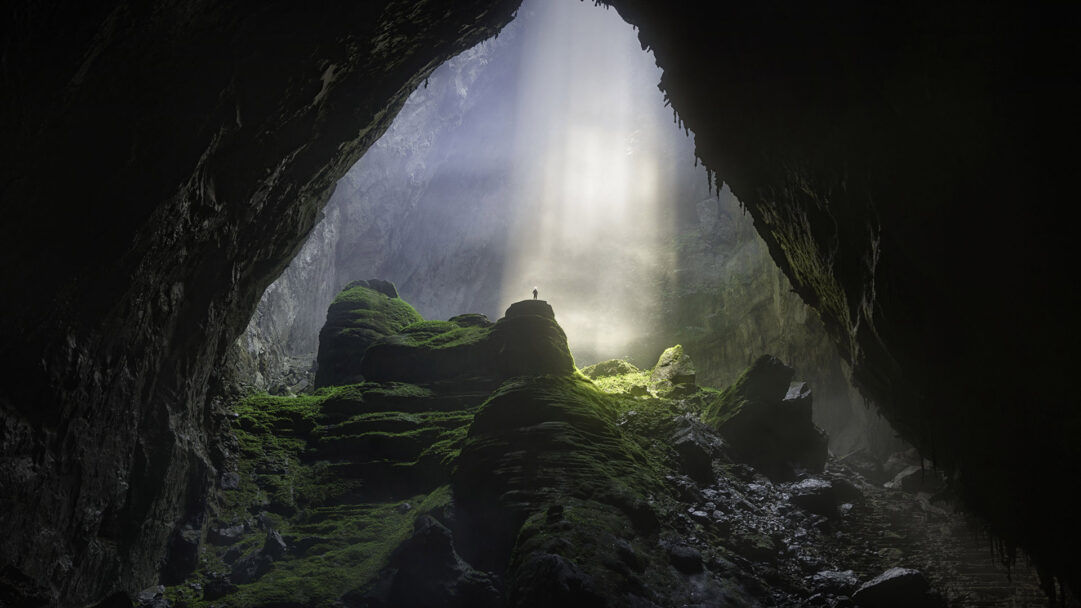 A breathtaking 4K wallpaper capturing the vast, moss-covered interior of Sơn Đoòng Cave in Vietnam, with a lone figure standing beneath a huge natural opening. Dramatic sunbeams pierce through the misty cavern opening, illuminating vibrant green moss on the colossal rock formations and creating an ethereal, awe-inspiring scene.