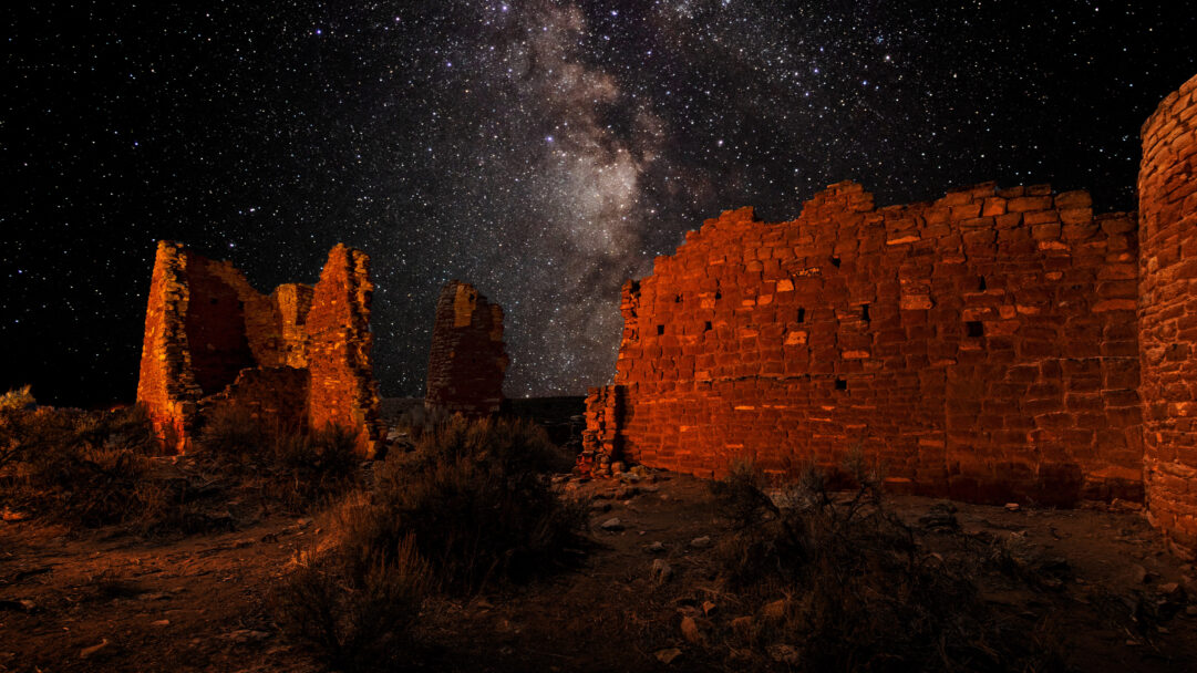 A captivating 4K wallpaper unveils the Square Tower Group at Hovenweep National Monument under a magnificent Milky Way night sky. The ancient stone structures glow with a warm, reddish light, providing a striking earthly contrast to the countless stars and luminous gas clouds of the Milky Way stretching across the dark expanse above.