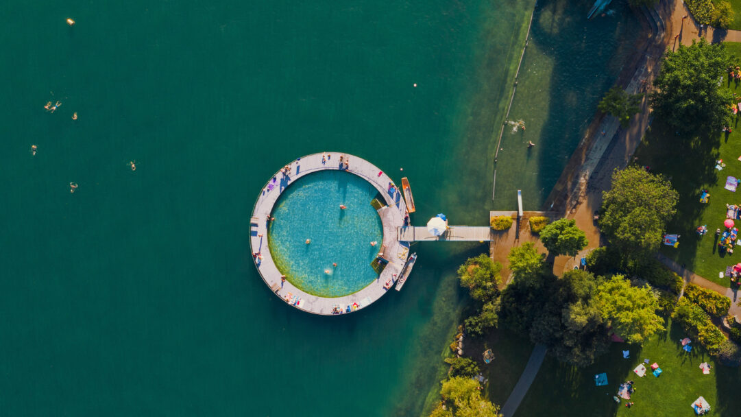 An expansive 4K wallpaper provides an aerial perspective of the Strandbad Tiefenbrunnen public pool on Lake Zurich, with its distinctive circular swimming area extending into the deep green lake. The image perfectly captures the vibrant summer atmosphere as numerous people swim, sunbathe on the surrounding wooden deck, and relax on the lush green parkland shoreline.