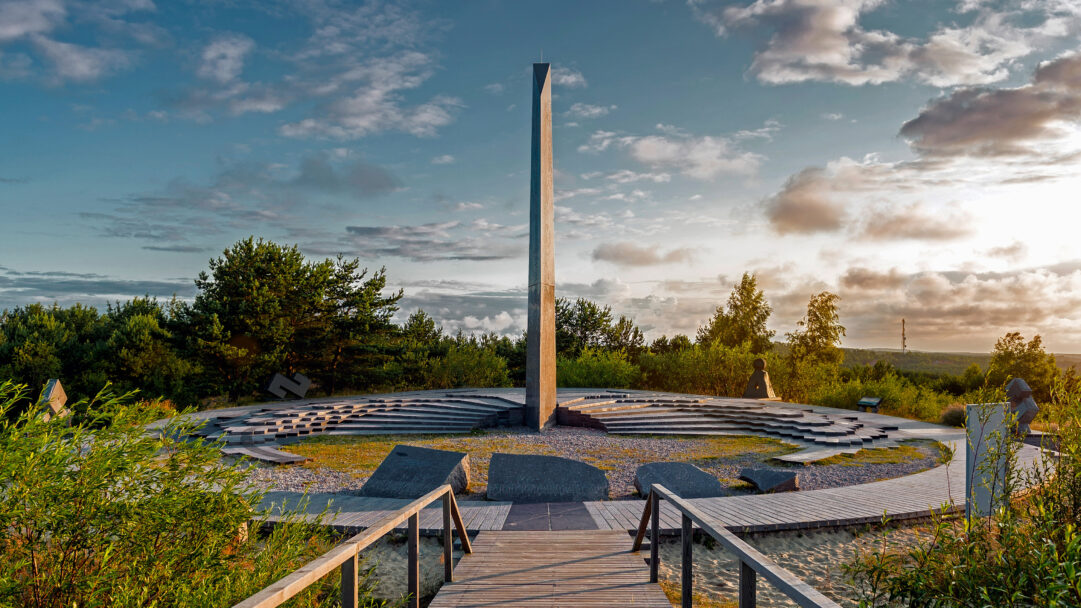 A magnificent 4K wallpaper presenting the monumental Sundial on Parnidis Dune in the Curonian Spit, Lithuania, amidst a vast green landscape under a partly cloudy sky. Golden sunlight bathes the scene, highlighting the sundial's stark form and the tranquil beauty of the natural surroundings.
