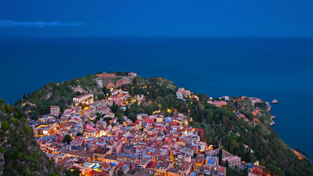An enchanting 4K wallpaper reveals the stunning night cityscape of Taormina, Sicily, nestled along the Italian coast. Warm golden lights from the buildings illuminate the terraced town, contrasting beautifully with the deep twilight blue of the Mediterranean Sea and sky above.