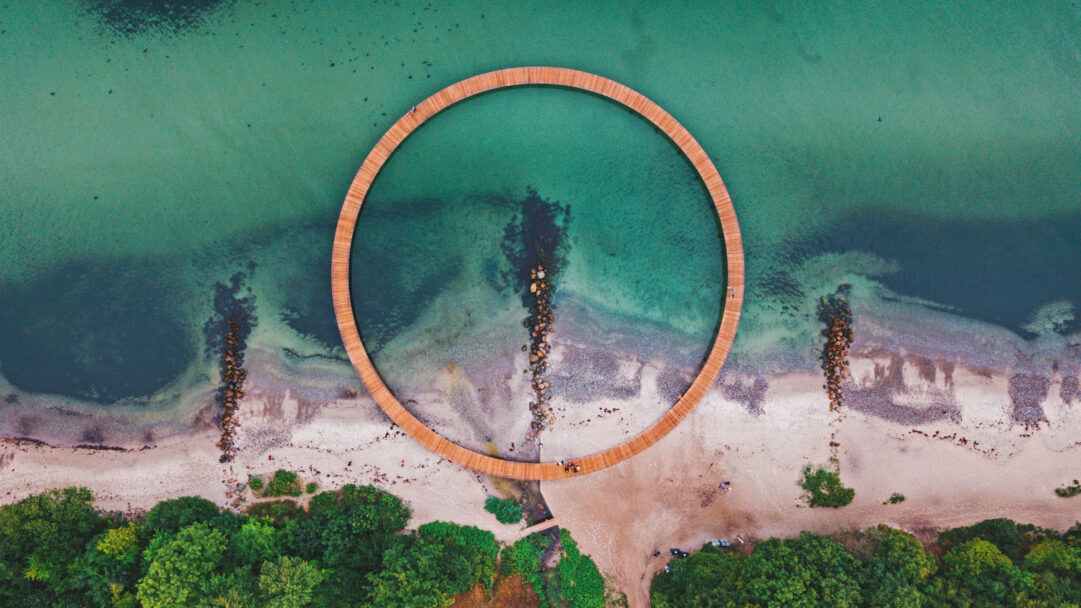 A captivating 4K wallpaper showcasing The Infinite Bridge in Aarhus, Denmark, as seen from an aerial perspective where the circular wooden walkway connects sandy beach and tranquil green waters. The striking interplay of the bridge's smooth curve against the varied textures of sand, sea, and scattered beachgoers creates a sense of serene, unending exploration.