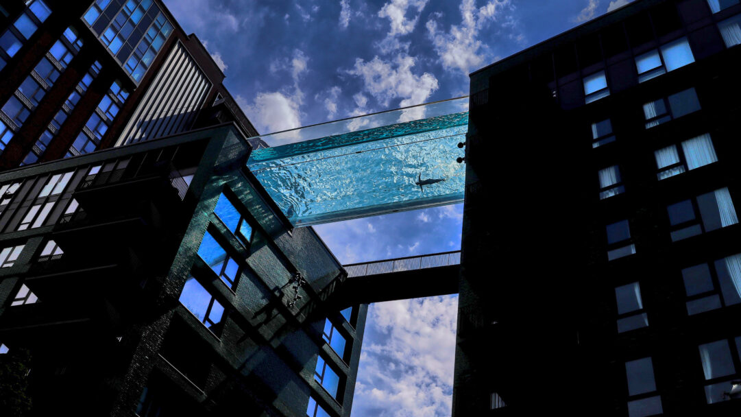 A mesmerizing 4K wallpaper of the Sky Pool at Embassy Gardens, London, England, an architectural marvel connecting two contemporary high-rise buildings under a bright, partly cloudy sky. A lone swimmer’s dark silhouette is visible through the clear, rippling turquoise water, creating a stunning visual contrast with the buildings' dark facades and the bright sky above.
