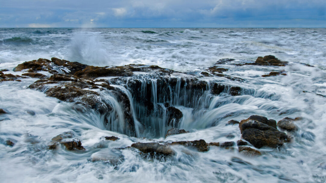 A mesmerizing 4K wallpaper showcasing Thor's Well on the rugged Cape Perpetua of the Oregon Coast, where powerful ocean waves crash and recede. Long exposure captures the frothy white waves dramatically draining into the deep, dark well, creating a powerful and ethereal natural spectacle under a brooding sky.