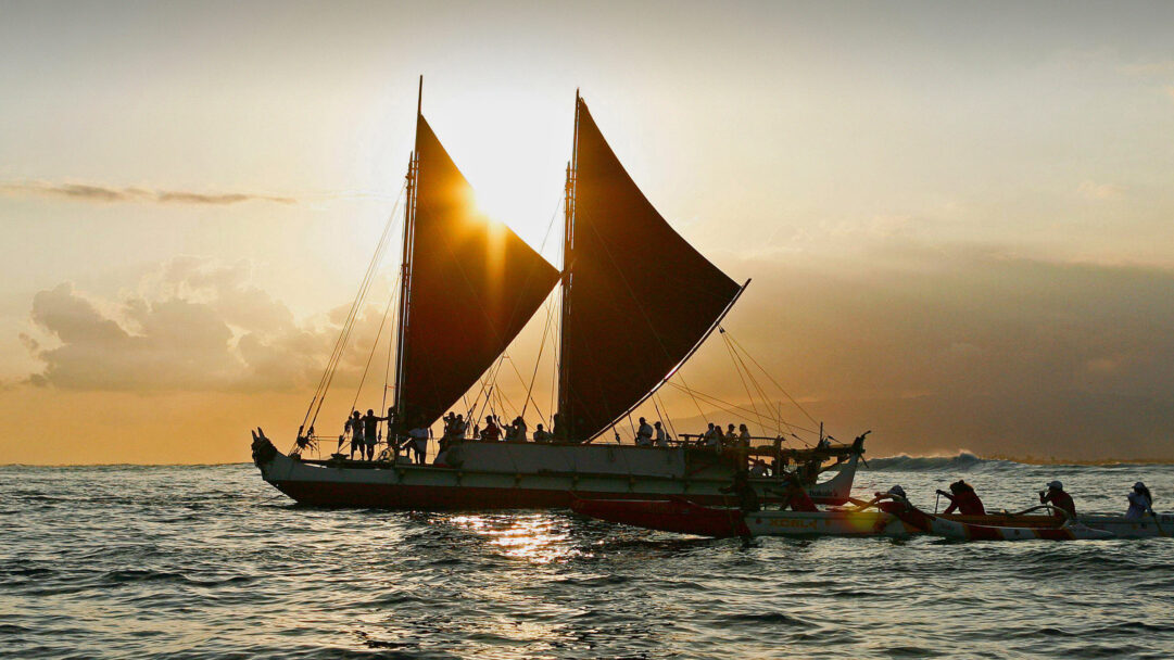An evocative 4K wallpaper portrays a traditional Hawaiian voyaging canoe sailing majestically on the open ocean as the sun dips below the horizon, with several outrigger canoes accompanying it. The brilliant golden sunset casts the canoe's distinctive sails and the numerous crew members into dramatic silhouette, creating a powerful scene of heritage and journey.