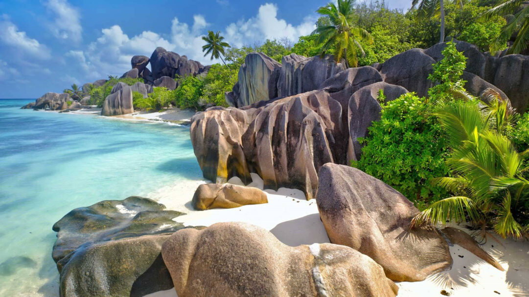 A captivating 4K wallpaper showcasing a pristine tropical beach, characterized by massive, weathered granite boulders scattered along its shore. The incredibly clear, vivid turquoise water gently laps against the white sand and the unique textures of the smooth rocks, creating a serene and inviting scene.
