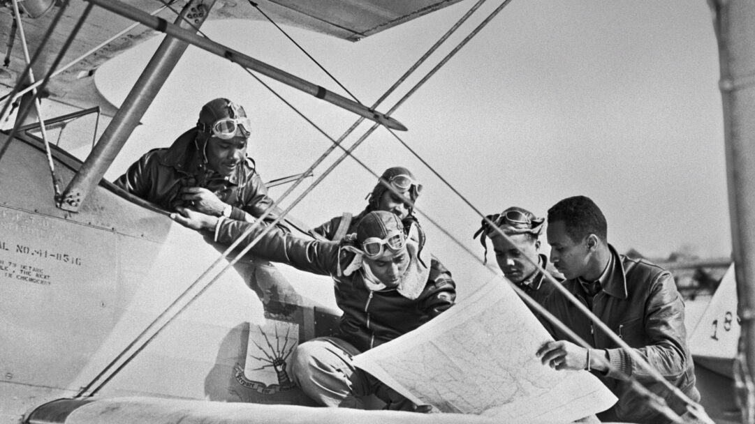A captivating 4K wallpaper showcasing four Tuskegee Airmen, dressed in flight helmets and leather jackets, intently studying a large map while gathered around the wing and fuselage of a vintage biplane. The classic black and white aesthetic highlights their focused expressions and the intricate details of the map, underscoring their shared resolve and the era's pioneering spirit.