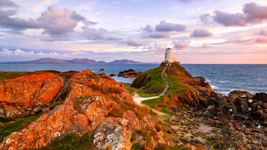 A breathtaking 4K wallpaper depicting Tŵr Mawr Lighthouse on Llanddwyn Island, Anglesey, Wales, nestled within a dramatic coastal landscape at sunset. The warm golden light of the sunset vividly illuminates the rugged orange-lichen-covered rocks and the winding path leading to the white lighthouse, creating a striking interplay against the soft pastel clouds and the deep blue sea.