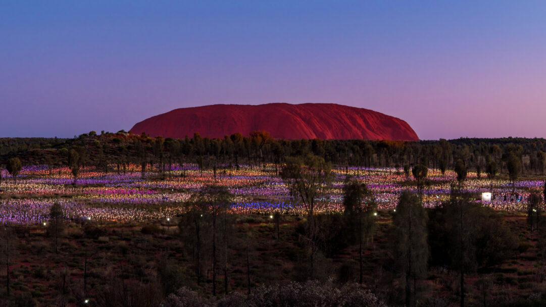 An enchanting 4K wallpaper capturing Uluru standing majestically against a twilight sky, overseeing the sprawling Field of Light art installation in the desert. Thousands of shimmering, multicolored lights create a mesmerizing carpet across the desert floor, reflecting the warm hues of the fading dusk and casting a magical glow.