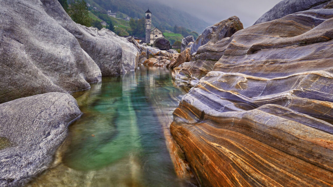 A captivating 4K wallpaper showcasing the Verzasca River flowing through a dramatic gorge of sculpted rocks in Lavertezzo, with a charming stone church nestled on the green hillside above. The remarkably clear, emerald-green water of the river contrasts beautifully with the intricate geological striations of the surrounding ancient rocks.