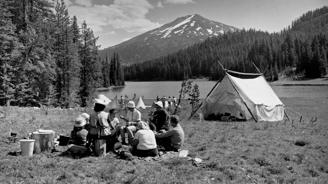 A captivating 4K wallpaper showcasing vintage Girl Scouts camping at Todd Lake in 1960, gathered in a grassy clearing. The black and white scene captures their outdoor camaraderie against a backdrop of dense forest, a calm mountain lake, and a majestic, snow-capped peak, imparting a timeless sense of adventure.