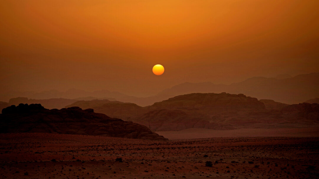 A captivating 4K wallpaper depicting the majestic Wadi Rum desert in Jordan at sunset, with a vast landscape of rocky mountains and sandy plains. The vibrant orange sun, hanging low in the hazy, warm sky, casts an intense, monochromatic glow over the layered, silhouetted terrain, creating a serene and dramatic mood.