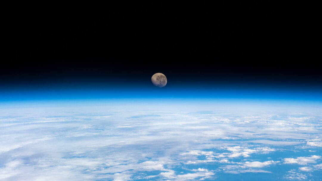 A cosmic 4K wallpaper features a detailed waning gibbous Moon suspended above the gentle curve of Earth's horizon, observed from the vast expanse of space. The dramatic transition from the pale blue atmospheric glow to the absolute blackness of space, alongside the Moon's textured surface, evokes a serene yet immense scale.