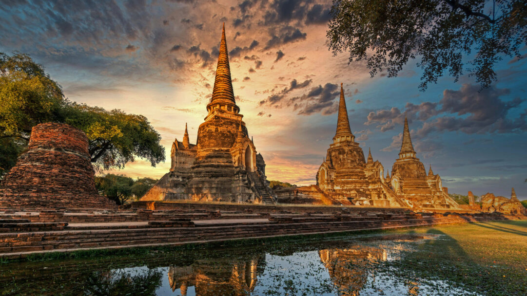 A magnificent 4K wallpaper presents the ancient ruins of Wat Phra Si Sanphet within Ayutthaya Historical Park, featuring multiple golden-hued chedis and structures. Golden light from the sunset dramatically illuminates the towering temple elements, casting their striking reflections across a tranquil foreground pool, evoking a sense of timeless grandeur.