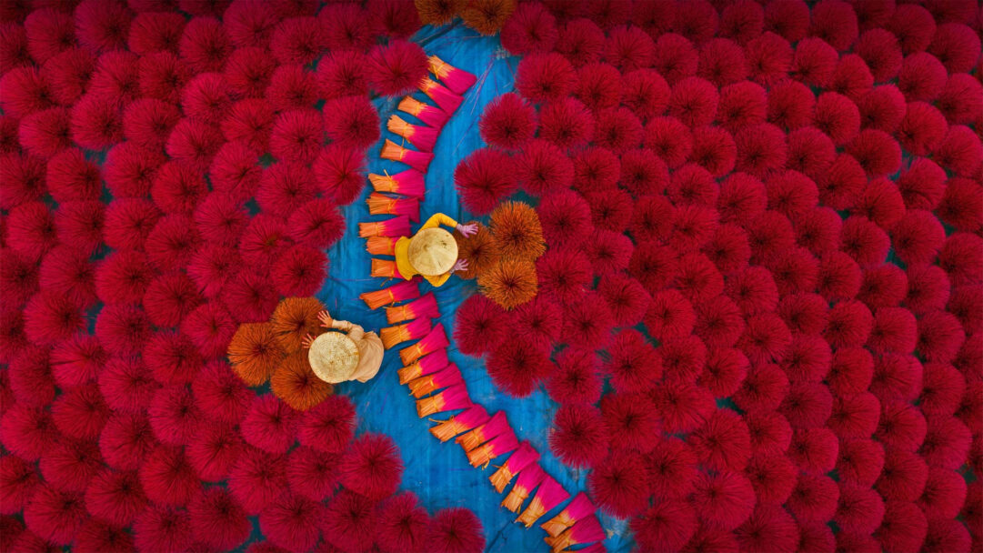 A breathtaking 4K wallpaper showcasing workers meticulously sorting vibrant red incense sticks in Huyện Ứng Hòa, Hanoi Vietnam. The aerial view emphasizes the stunning contrast of crimson incense bundles fanned out across a vivid blue drying tarp, creating a striking mosaic of color and texture.